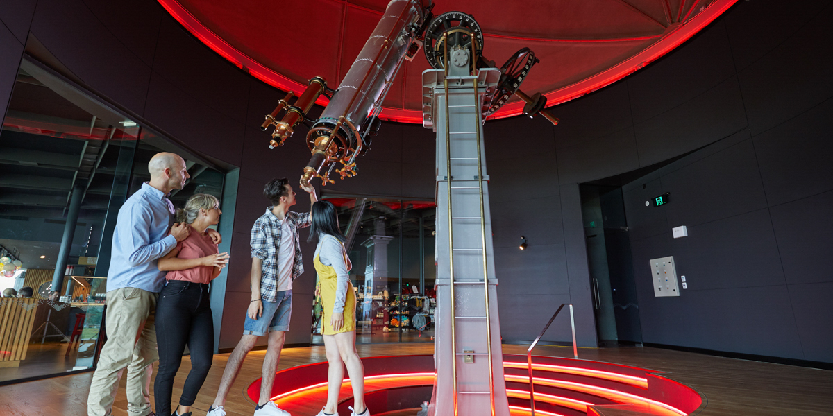 The image shows a group of four people standing inside a modern observatory, gathered around a large telescope. The telescope is mounted on a central pedestal, and a red glowing circular light at the base of the structure creates an atmospheric ambiance.