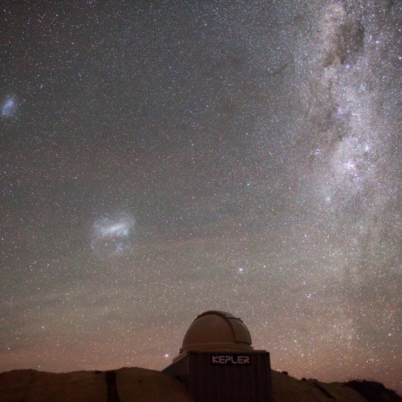 The image shows a nighttime sky filled with a vast array of stars and cosmic formations, including the bright, cloudy appearance of the Milky Way galaxy stretching across the sky. Below the celestial scene is an observatory dome labeled "KEPLER," located 