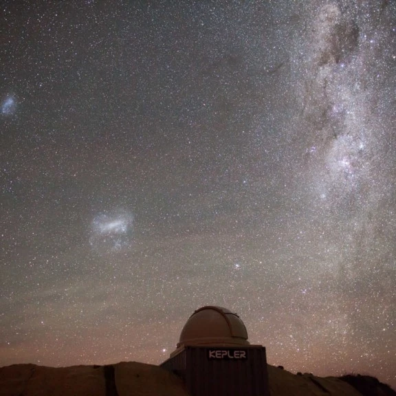 The image shows a nighttime sky filled with a vast array of stars and cosmic formations, including the bright, cloudy appearance of the Milky Way galaxy stretching across the sky. Below the celestial scene is an observatory dome labeled "KEPLER," located 