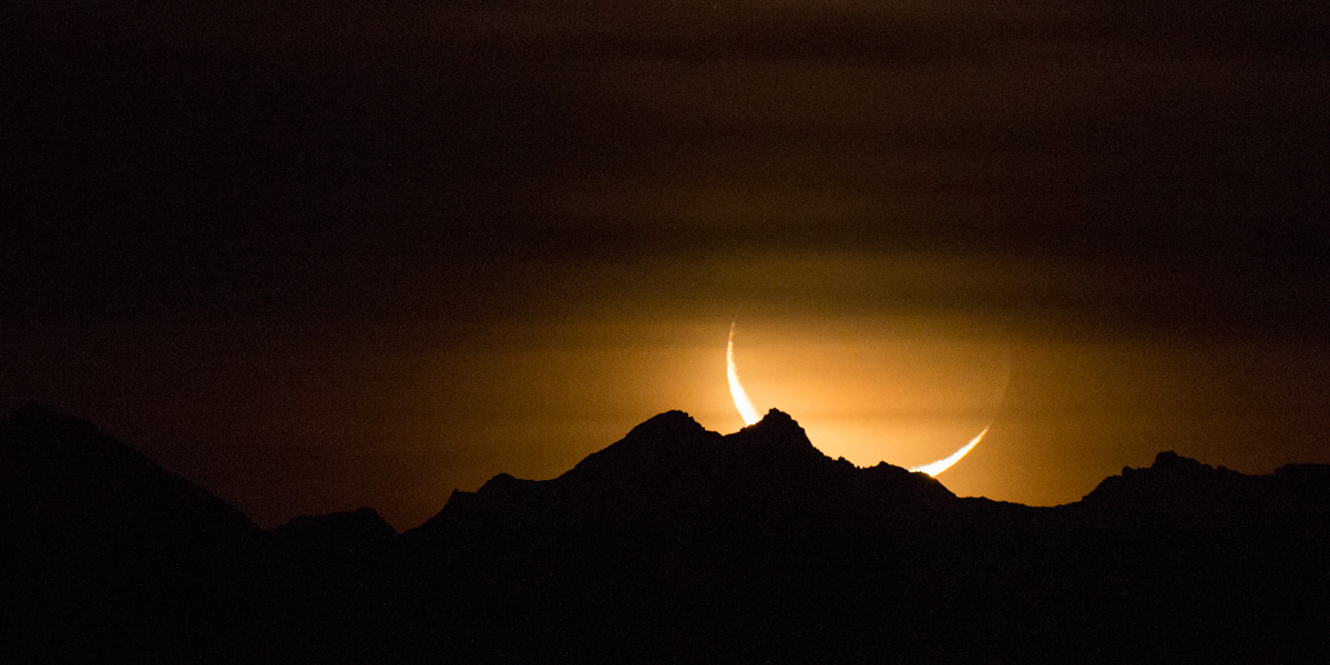 Crescent moon rising from behind mountain range at night