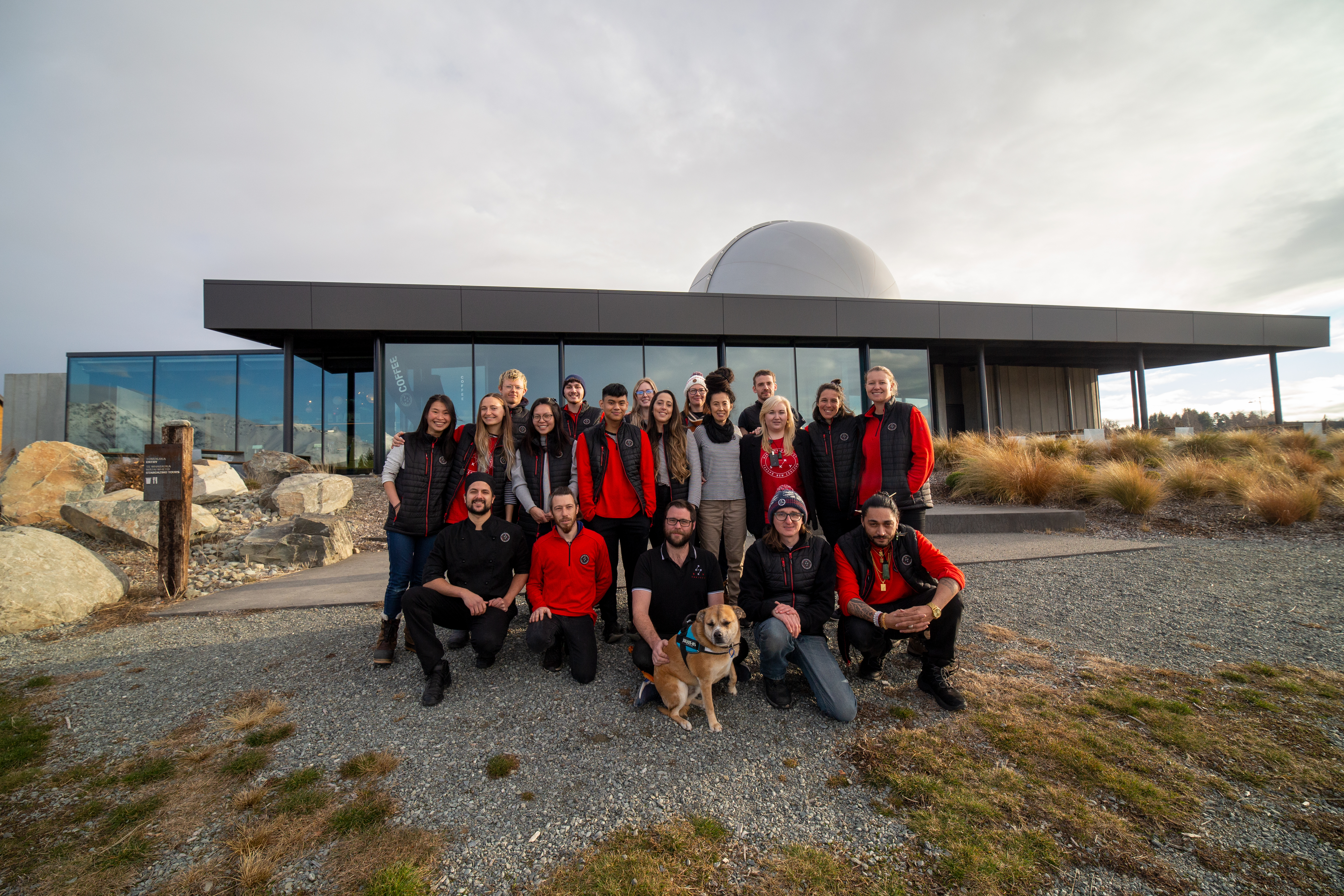 The image shows a group of people posing for a group photo in front of a modern building with large glass windows and a dome on the roof, likely an observatory. The group, dressed in casual outdoor clothing and red vests or jackets, is smiling and gather